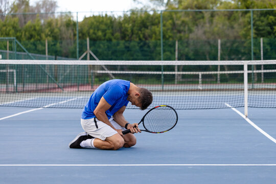 Male athlete kneeling on tennis court in blue shirt and white shorts holding racket under net