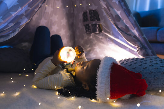 African American child wearing Santa-hat in lit star tent at home holding glowing orb, copy space