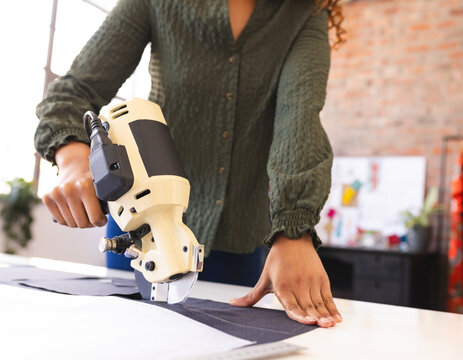 African American woman operating fabric cutter and steadying dark fabric on white table in studio