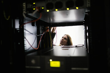 female IT technician reaching into server rack in server room inspecting red cables and LED lights