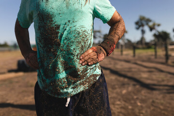 Man standing outdoors in bright sun on obstacle course with rope drawstring, wrist wraps and mud