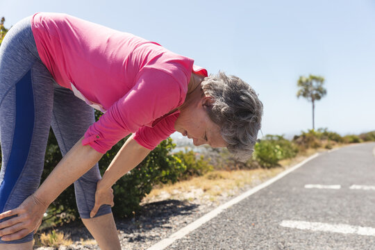 Mature woman leaning forward catching breath on paved path in pink top and grey-blue leggings
