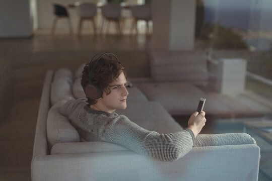 Man in his twenties sitting on sofa wearing headphones holding smartphone in open plan living space