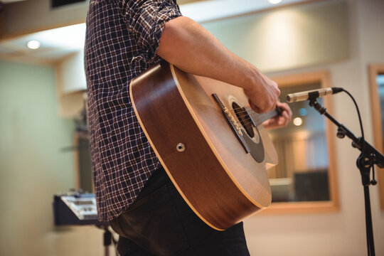 Male musician in dark plaid shirt playing acoustic guitar by mic stand, console and studio window