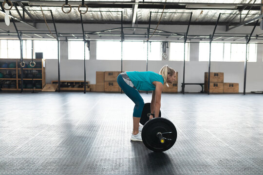 Female athlete wearing teal top and leggings gripping barbell on floor at CrossFit gym under rings