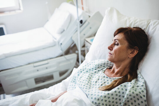Senior woman lying on hospital bed in gown with pillow-blanket beside bed IV pole, copy space