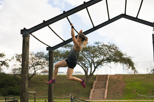 Mid adult woman hanging on metal monkey bars at outdoor fitness area with wooden staircase, tree