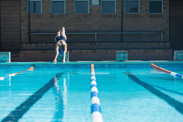 Female competitive swimmer diving off starting block into outdoor lap pool in swimsuit, goggles