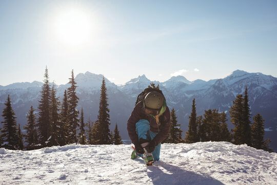 Skier kneeling on snow ridge adjusting boots, wearing jacket, helmet and goggles under bright sun