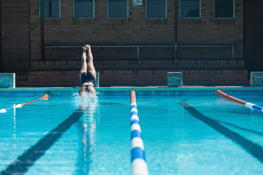 Female swimmer in dark swimsuit diving headfirst into water at pool deck with colored lane ropes