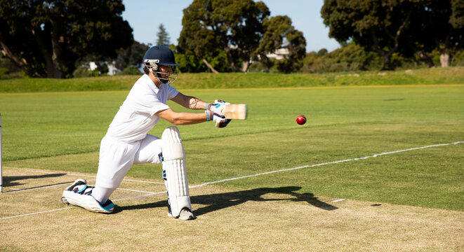 Male cricketer wearing helmet kneeling at crease swinging bat toward ball near stumps, copy space