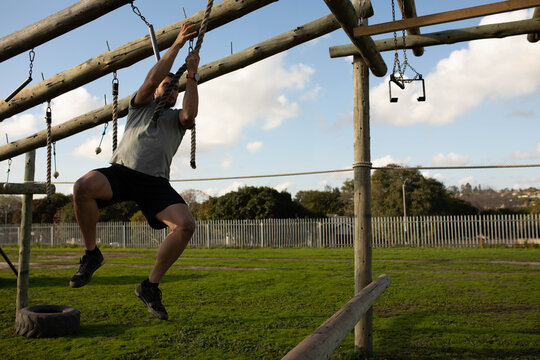 Male athlete grabbing rope, swinging to bar above tire on obstacle course on grass, copy space