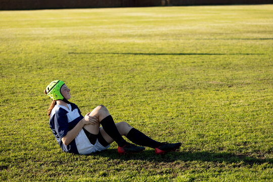 Teenage girl sitting on grass field holding knee, wearing scrum cap and rugby kit, copy space