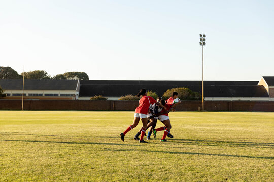 Diverse teenage female rugby teammates gripping rugby ball while tackling opponent on school pitch