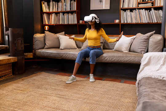 African American woman using virtual reality headset on sofa with pillows in living room library