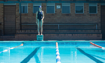 Female swimmer standing atop starting block at lap pool wearing navy swimsuit, swim cap and goggles