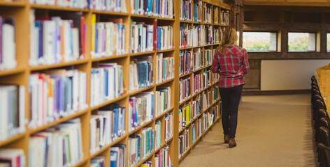 Female student walking aisle between shelves and tables with chairs under windows, copy space