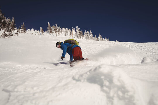Male snowboarder carving through deep powder on mountain slope with backpack under clear blue sky