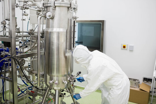 Woman technician in protective coverall adjusting valve on bioreactor in clean room, copy space