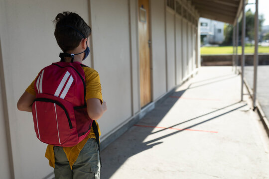 Child boy standing in school corridor wearing protective face mask and red backpack, copy space