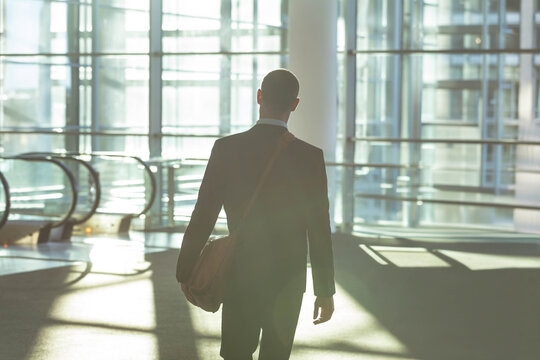 Male businessman walking through sunlit atrium with glass panels, carrying leather shoulder bag