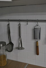 Three kitchen utensils hanging on a metal rail with hooks against a clean white tiled kitchen wall, with a glass jar of pasta visible in the background.