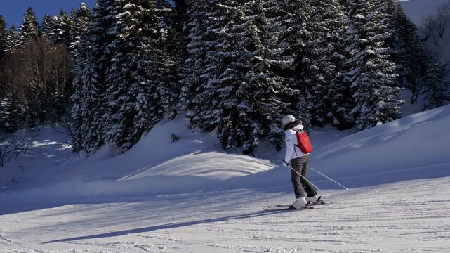 Lone skier moves silently across white snow piste. Skier makes journey over glistening winter terrain in ski resort. Skier skiing on snow downhill under expansive blue sky and verdant trees.