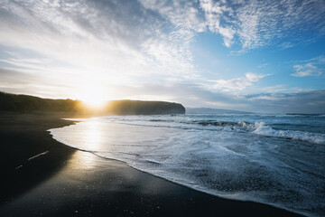 Dramatic sunset over black sand beach with ocean waves and sun rays through clouds