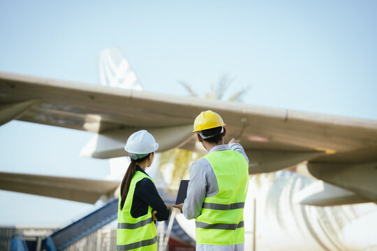 Two engineers in safety helmets and reflective vests inspect an aircraft landing gear tire while reviewing maintenance data on digital tablets at an airport maintenance area.
