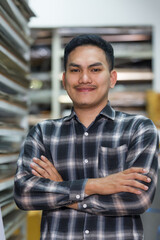 Obraz premium Asian adult man professional carpenter standing with crossed arms inside woodworking supplies shop representing skilled tradesman managing small business wood material inventory