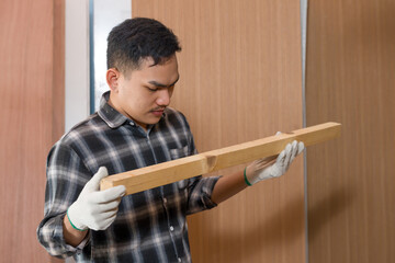 Asian male woodworking business owner holding wooden plank inspecting surface quality inside interior furniture material shop, professional carpenter checking straightness for furniture manufacturing