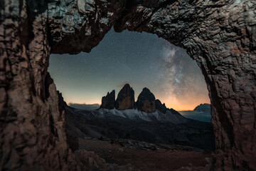 Fototapeta premium Rocky cave Grotta delle Tre Cime framing Tre Cime di Lavaredo mountain peaks with Milky Way in Dolomites, Italy