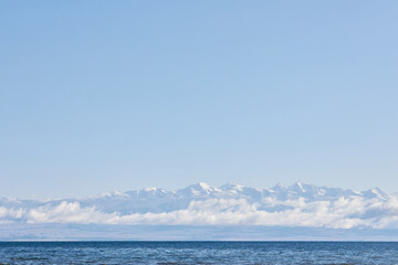 Fototapeta premium Calm blue ocean with distant snow-capped mountain range under clear sky. Serene nature scene for travel destination advertising and background use.
