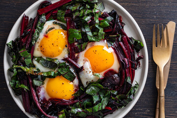 A vibrant, high-angle close-up photograph of three sunny-side up eggs fried with a generous helping of red beet stems and dark green leaves in a white plate, served with golden cutlery on a dark woode