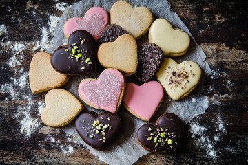 Rustic, heart cookies decorated with pink sugar and crushed pistachios on a sheet of weathered paper. The background is a dark, grainy wood table.
