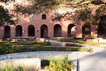 Brick building with arched doorways, lantern-style lights face courtyard of gravel beds, stone benches, winding paths. Tree-filtered sunlight adds depth, visual rhythm, college campus, Waco, TX