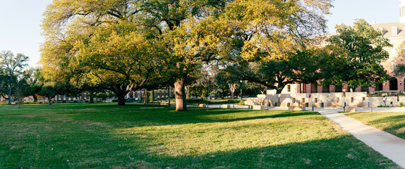 Panorama view red-brick building with arched windows with tower rises behind shaded lawn, square-cut stone wall. Benches and walkways complete peaceful, well-maintained campus scene, Waco, TX