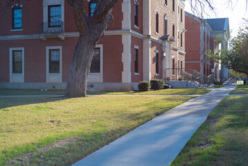 Red-brick academic building with white columns, walkway and balanced windows overlooks quiet lawn. Tree shadows and soft sunlight evoke a timeless, scholarly college campus setting in Waco, Texas