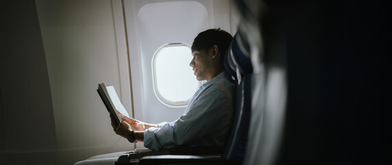 Asian businessman reading a book while sitting by the airplane window, representing calm in-flight time, business travel, and comfortable air transportation.