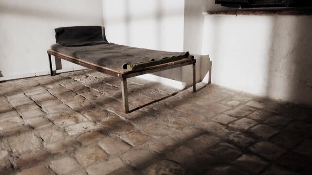 The camera caresses the stone floor, tilting slowly up, to reveal an authentic old prison bed. as the camera movement, shadows of the prison door are casting on the floor and walls. a 4K video clip.