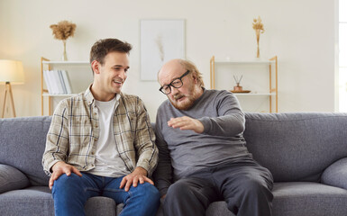 Senior father and adult son talking on couch at home. Happy young man listening to healthy...
