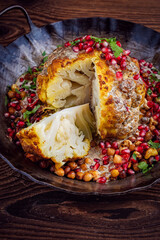 Vegetarian dish - whole oven baked roasted cauliflower with tahini sauce, chickpeas and pomegranate in rustic iron skillet pan served as close-up on a wooden board