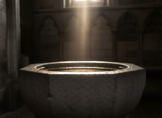 Stone baptismal font in a dark church with a ray of light illuminating the water inside. Religious tradition for baptism ceremony.