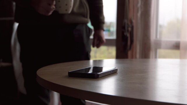 Nigra inteligenta telefono kuŝas sur la tablo. Viro kun trinkujo. Kampara domo. Endome. Proksima bildo. Sen vizaĝo. Aktoro. Ripozanta. Distanca laboro.