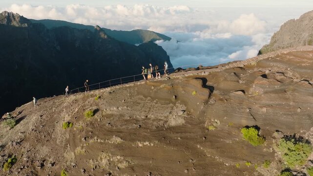 Cinematic side-view aerial drone shot capturing hikers walking along the exposed mountain ridge on famous trail from Pico do Arieiro to Pico Ruivo. The camera follows the ridge from the sunlit side 
