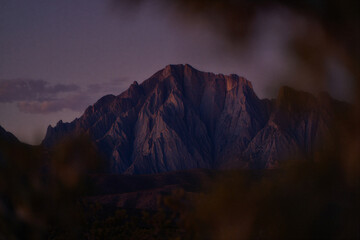 Majestic mountain peak illuminated by the soft glow of twilight with dramatic shadows and a hazy sky overhead viewed through foreground foliage