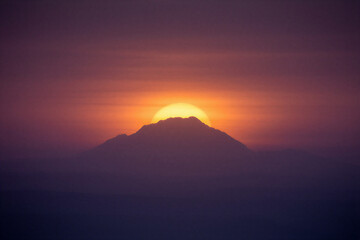 Majestic mountain silhouette against a vibrant sunset with sun glowing brightly behind the peak casting a warm orangepurple hue across the sky