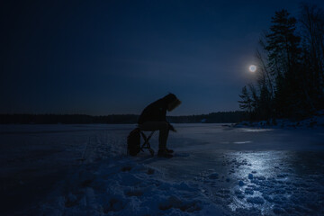 Silhouette of a person ice fishing on a frozen lake at night under moonlight, Finland.