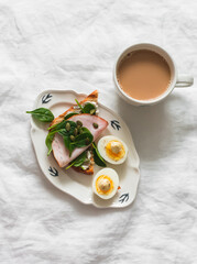 Delicious grilled breakfast - bread toast with cream cheese, spinach, ham, pumpkin seeds, boiled egg and coffee with milk on a light background, top view