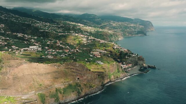 A smooth drone shot filmed near Cascata dos Anjos, capturing the rugged south coast of Madeira Island. The camera faces east along the coastline, revealing dramatic cliffs, ocean waves